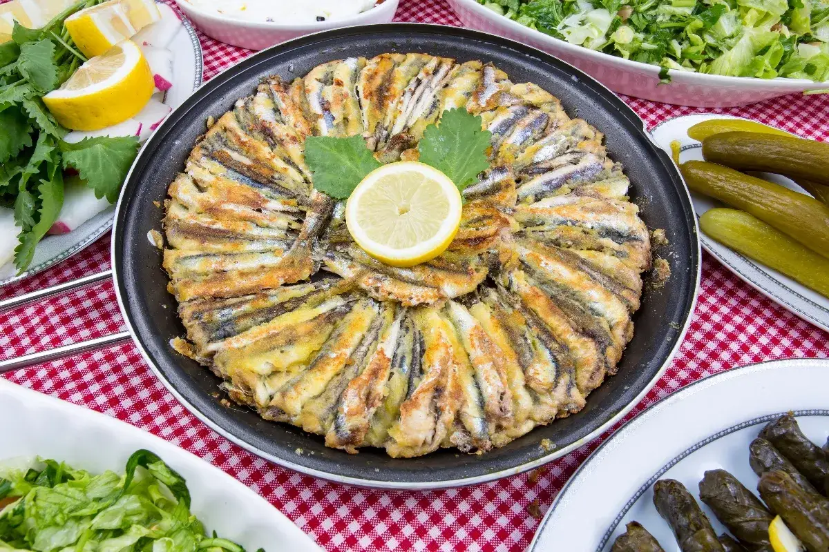 Parmigiana di alici dorate e croccanti, disposte a cerchio in una padella, con una fetta di limone al centro. Accompagnata da insalata e cetriolini.
