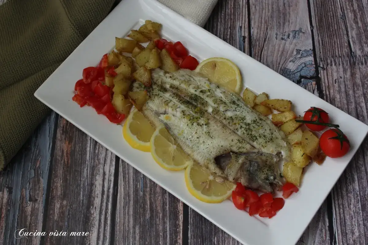 Sogliola al forno con patate arrosto, pomodorini e spicchi di limone su un piatto bianco.