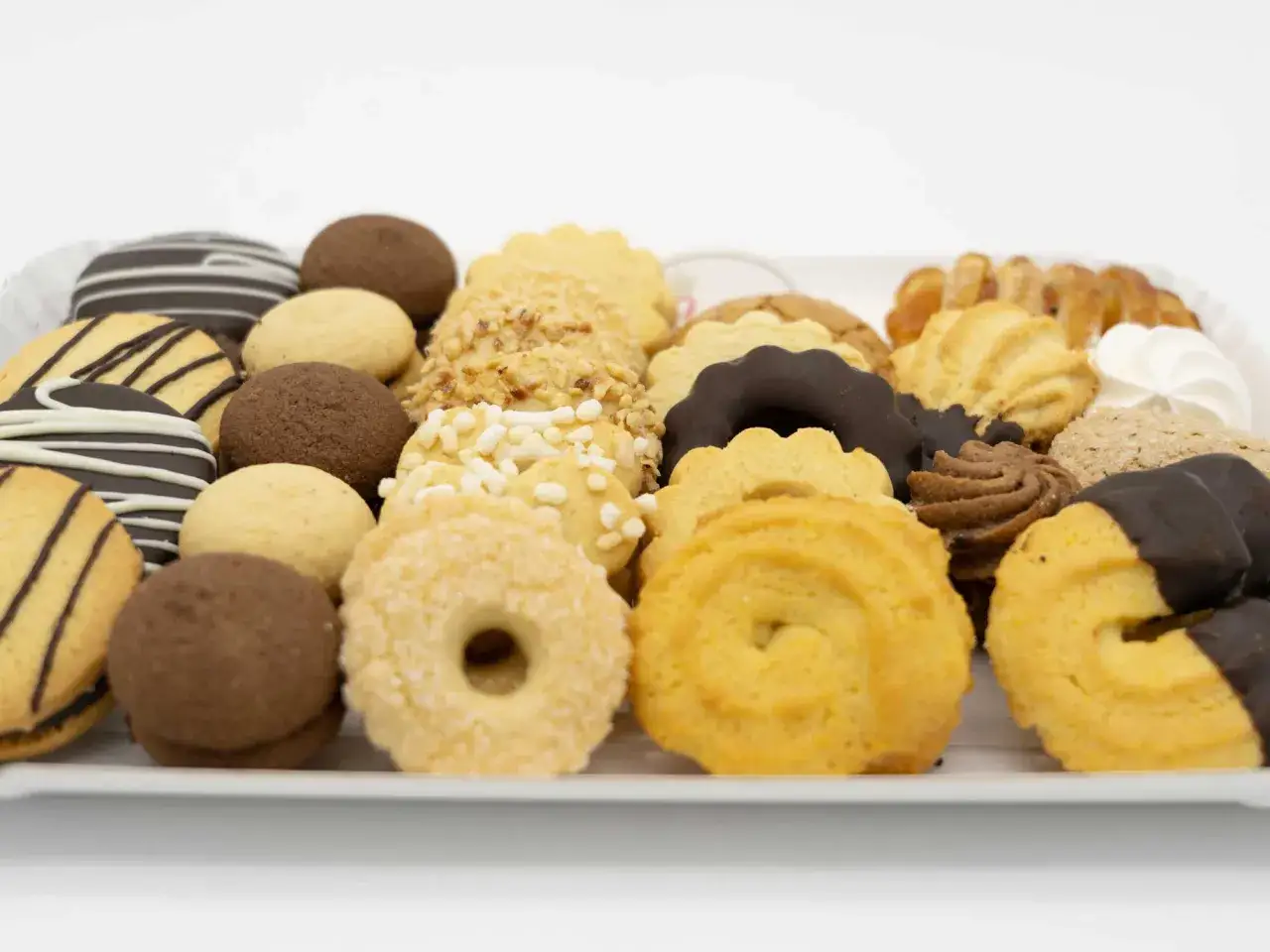 Vassoio pieno di biscotti assortiti, alcuni con glassa al cioccolato, altri con granella di nocciole e zuccherini. Tra questi, deliziosi biscotti con farina di riso, perfetti per ogni occasione.
