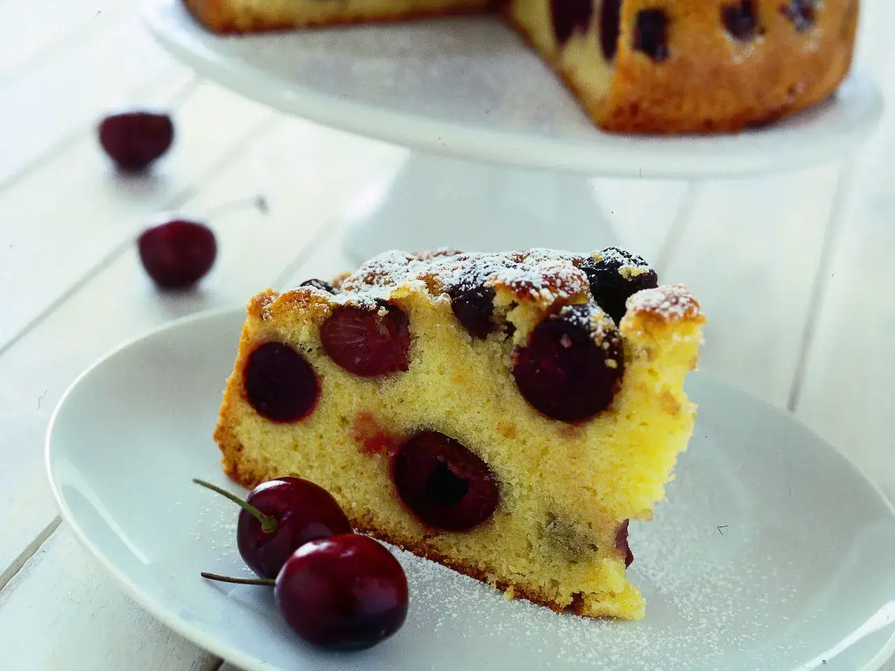 Una fetta di torta alle ciliegie, cosparsa di zucchero a velo, con ciliegie fresche sul piatto.