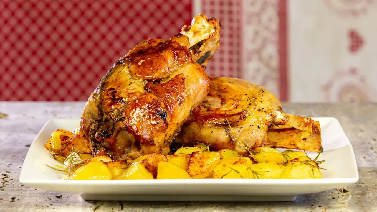 Due stinchi di maiale al forno dorati e succulenti, serviti con patate arrosto e rosmarino su un piatto bianco.