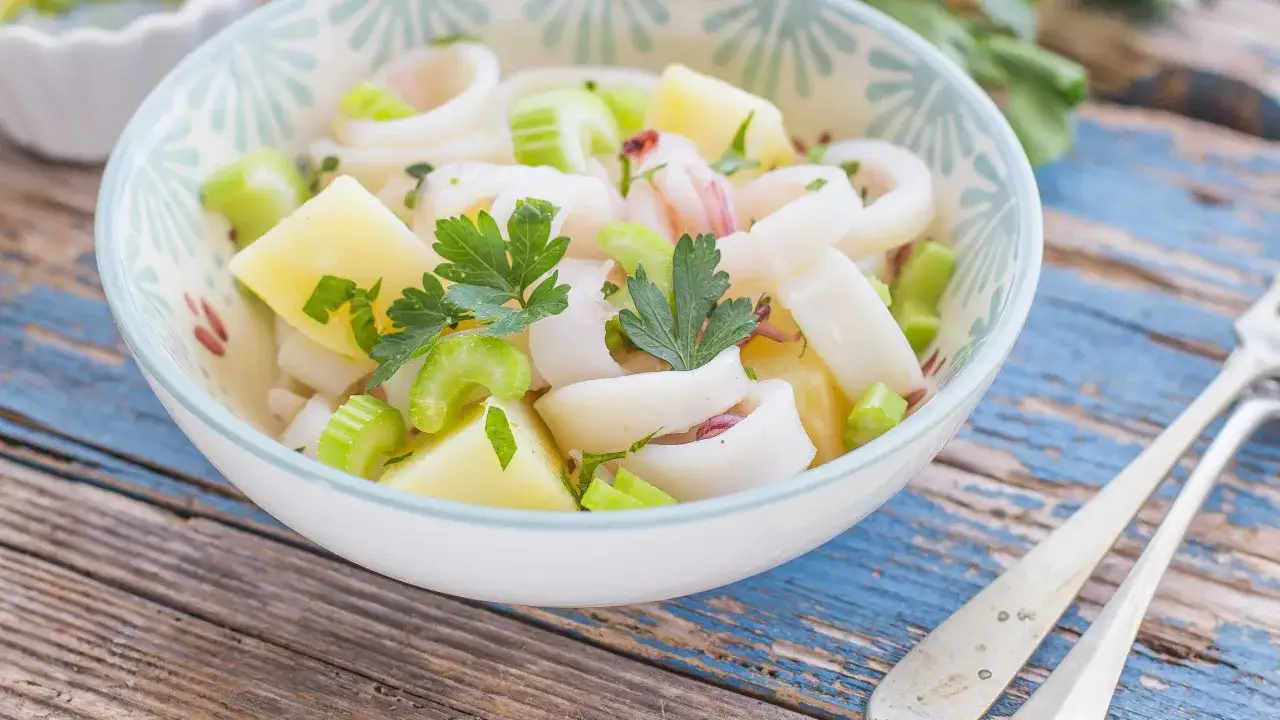 Insalata di calamari fresca con patate, sedano e prezzemolo, servita in una ciotola decorata su un tavolo di legno blu.