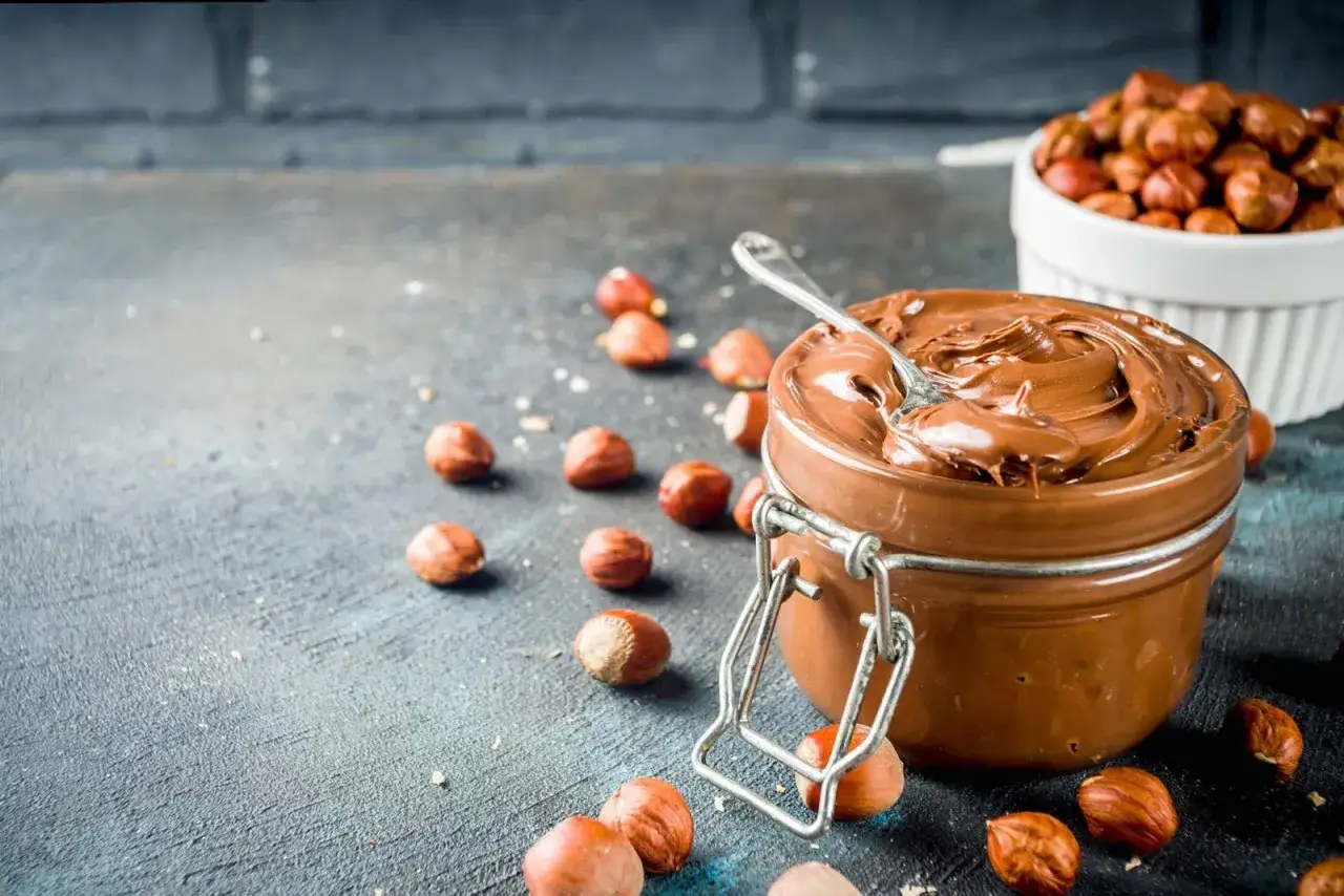 Crema di nocciole fatta in casa, cremosa e invitante, con nocciole sparse intorno. Un cucchiaino affonda nella delizia.
