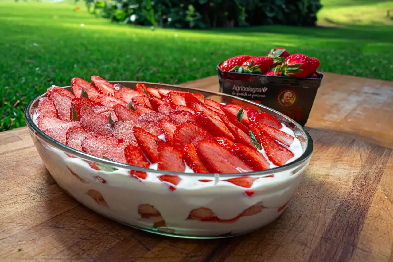 Un invitante tiramis&ugrave; alle fragole in una pirofila di vetro, guarnito con fragole fresche affettate e foglie verdi. Sullo sfondo, un prato verde e una vaschetta di fragole.
