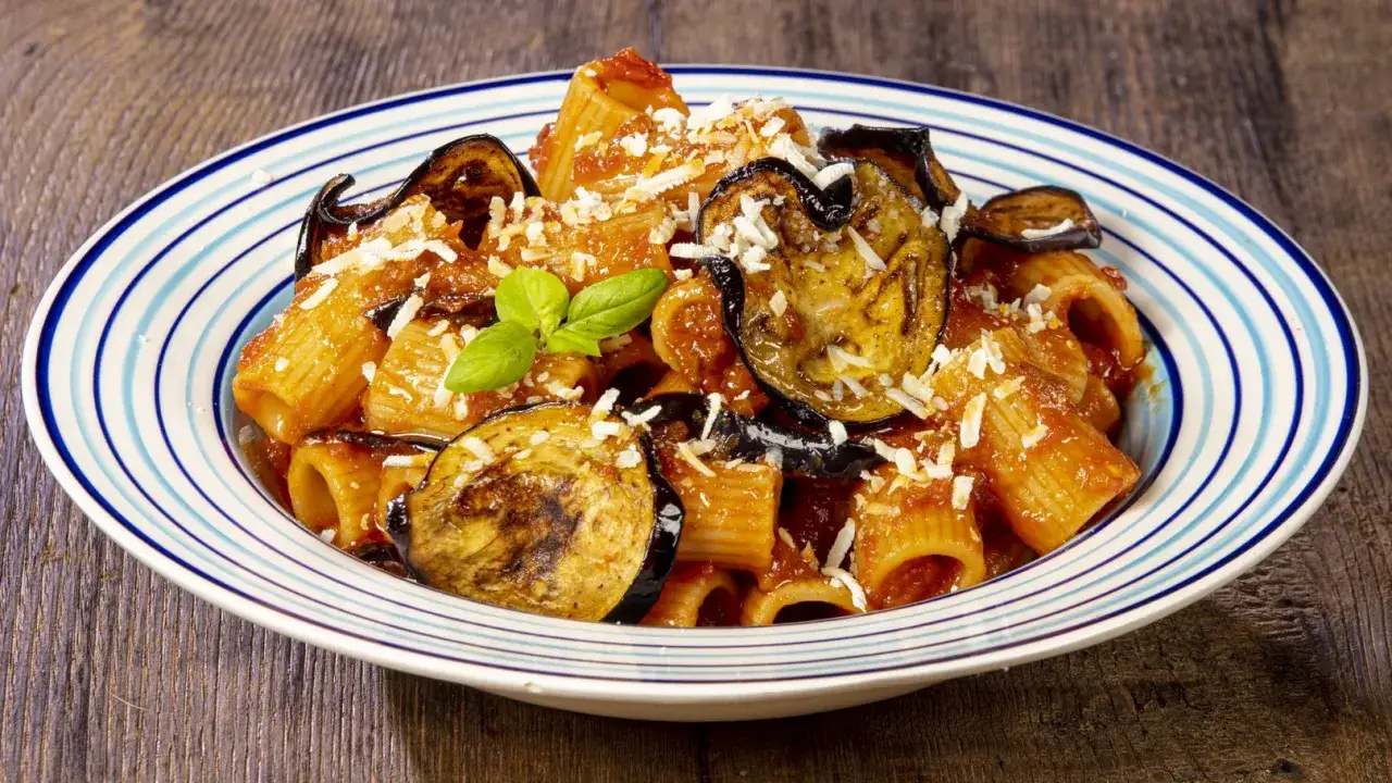 Un piatto di pasta alla norma con melanzane fritte, pomodoro e ricotta salata grattugiata, guarnito con basilico fresco.