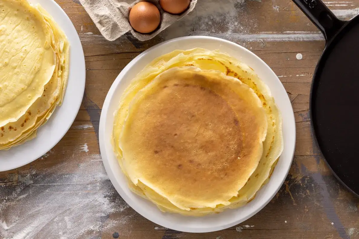 Piatto di crepes senza burro, dorate e leggere, pronte per essere farcite. Accanto, uova e una padella per la preparazione.