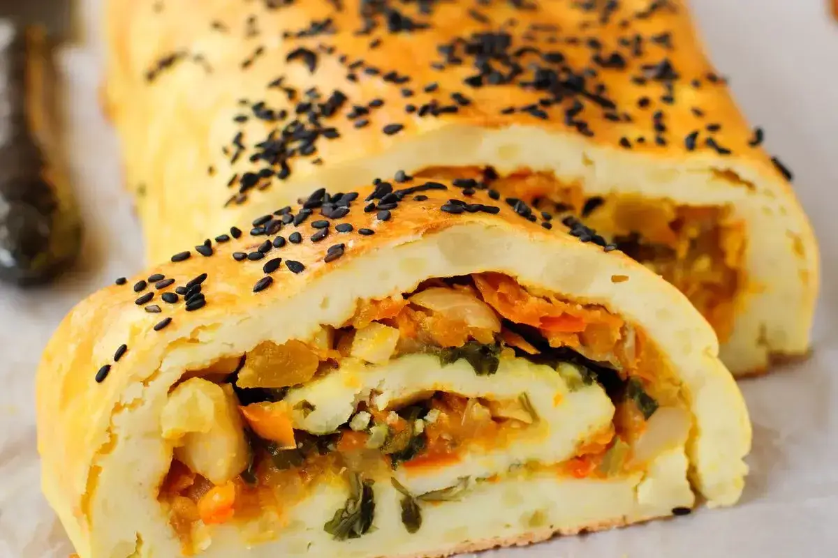 Rotolo di patate dorato, cosparso di semi neri, con un ripieno colorato di verdure.