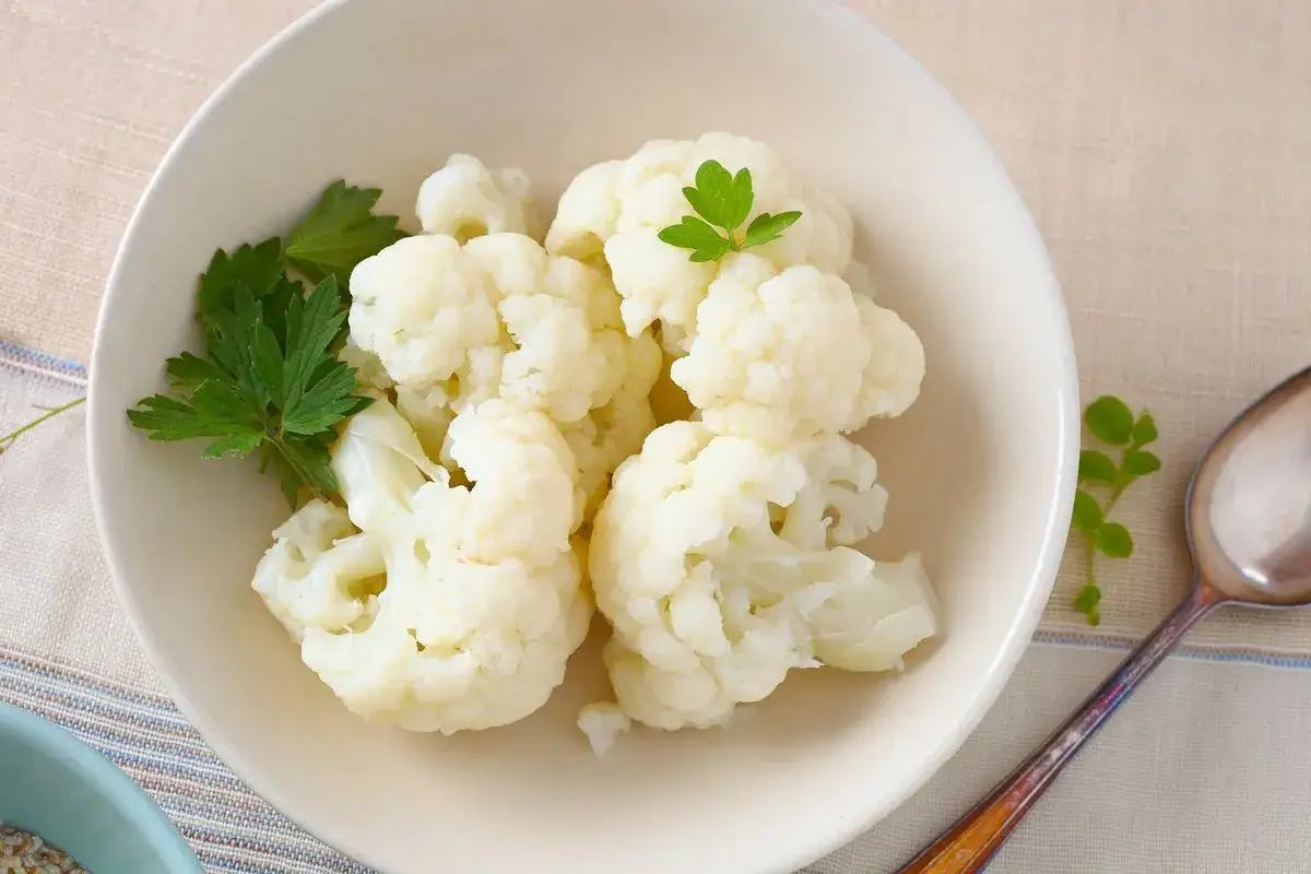 Cavolfiore bollito servito in una ciotola bianca, guarnito con prezzemolo fresco. Un cucchiaio d'argento &egrave; accanto.