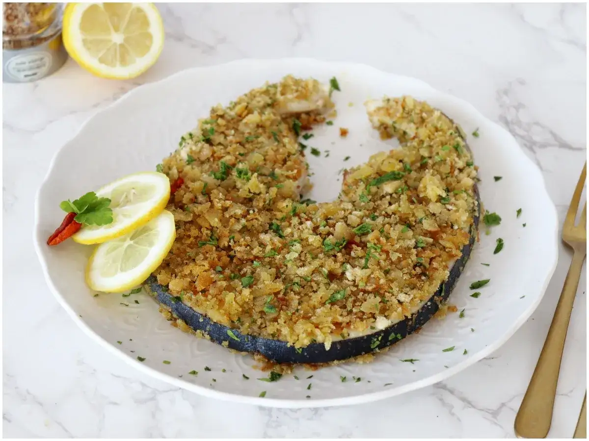 Pesce spada alla palermitana, impanato e cotto al forno, servito con limone e peperoncino su un piatto bianco.