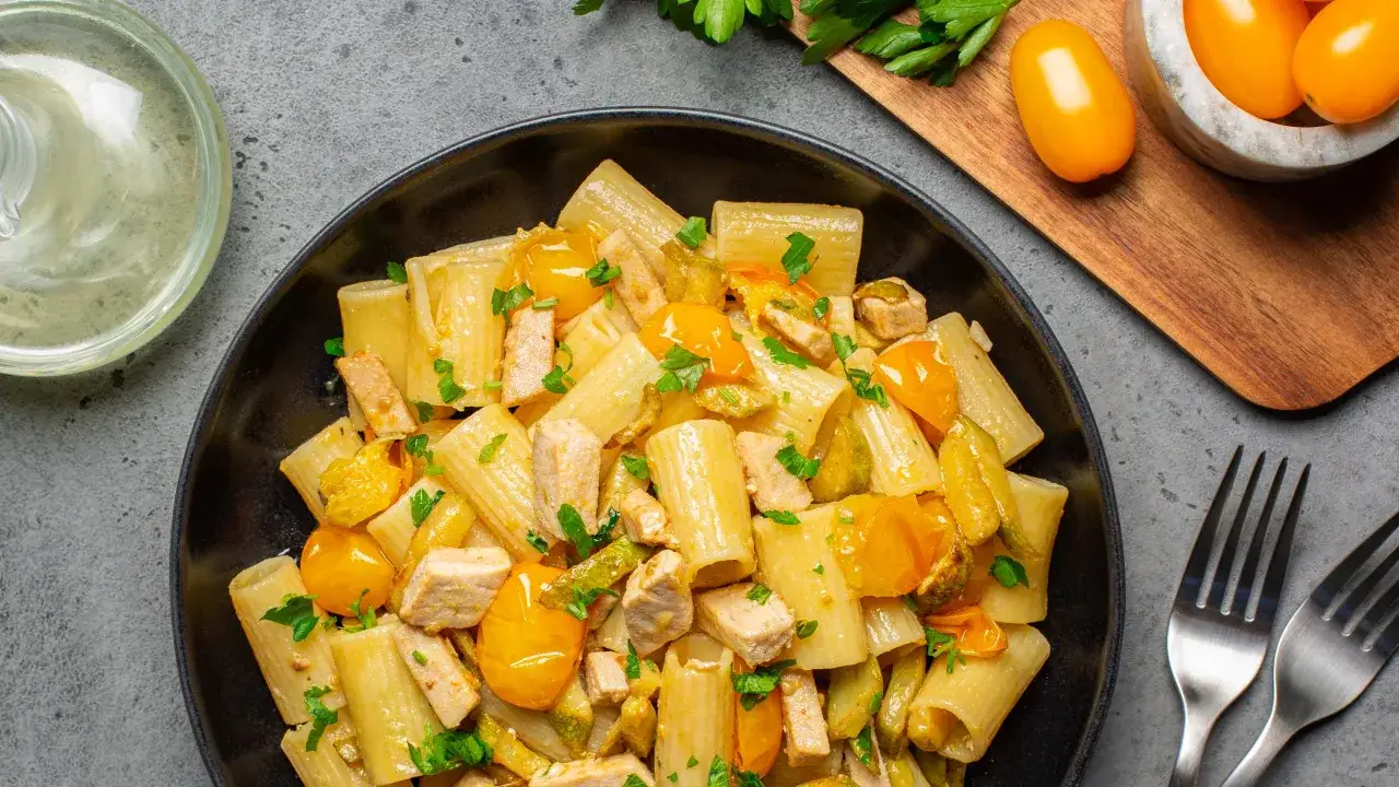Piatto di rigatoni conditi con cubetti di pollo, zucchine e succosi pomodorini gialli, guarnito con prezzemolo fresco.