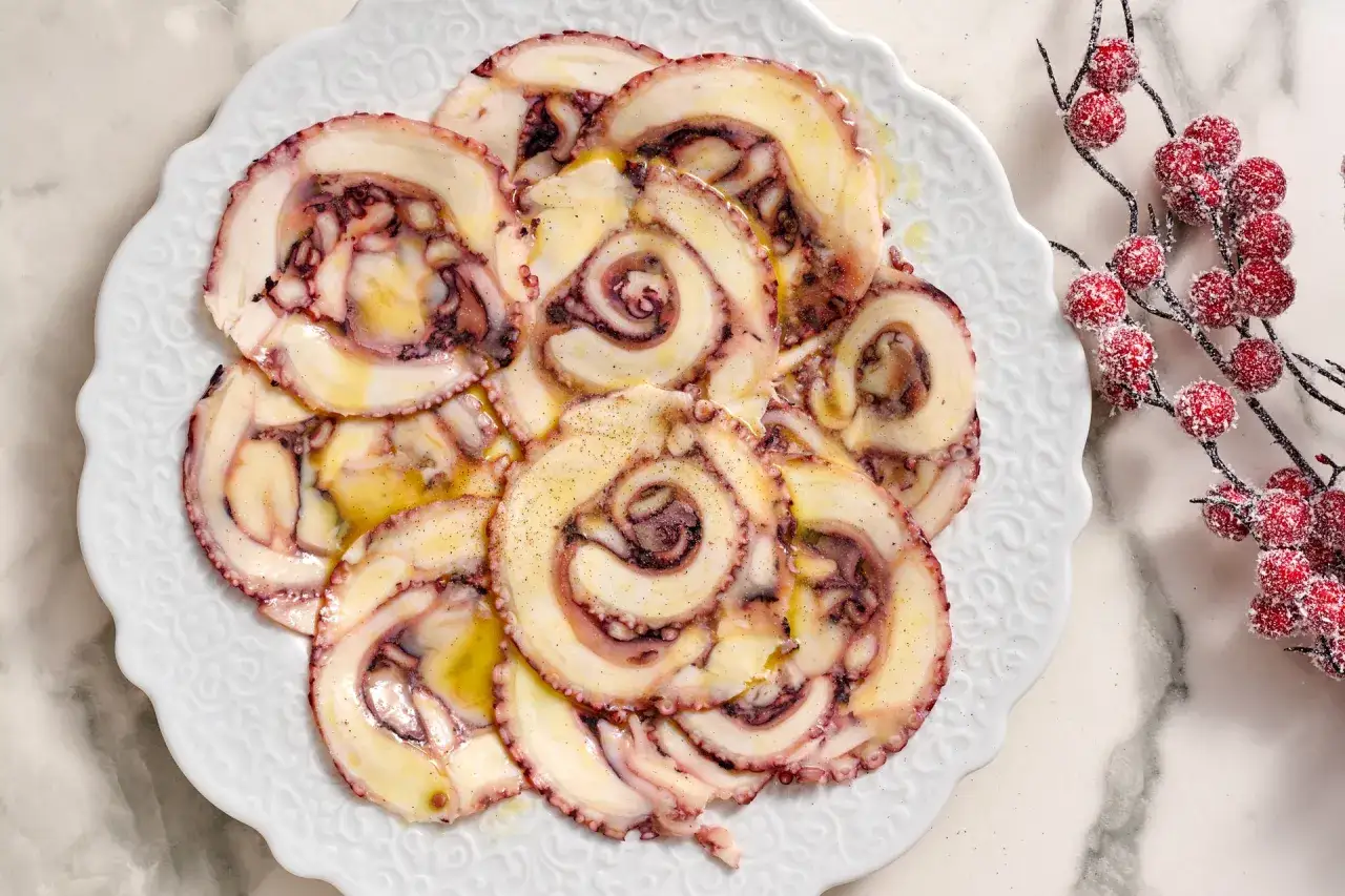 Fette di carpaccio di polpo condite con olio e pepe su un piatto bianco decorato.
