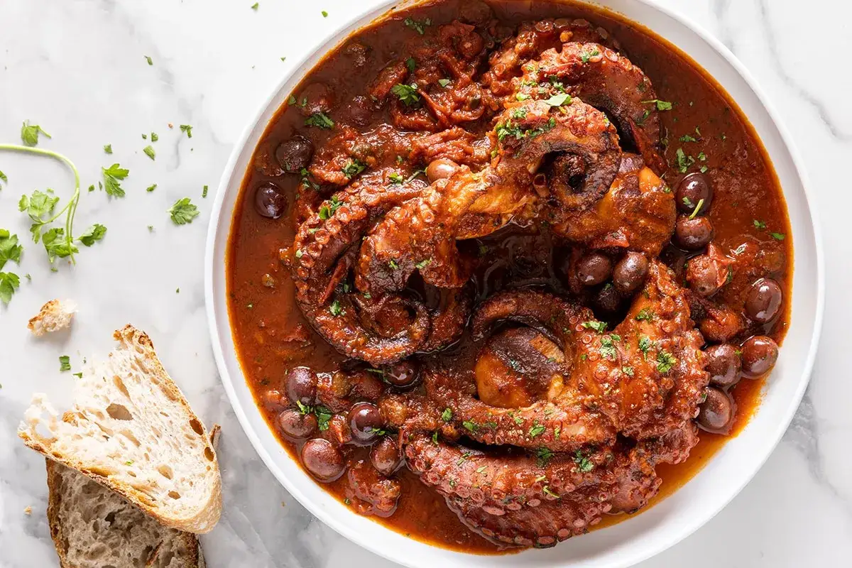 Un saporito polpo alla luciana in umido con olive scure, guarnito con prezzemolo fresco e accompagnato da fette di pane rustico.