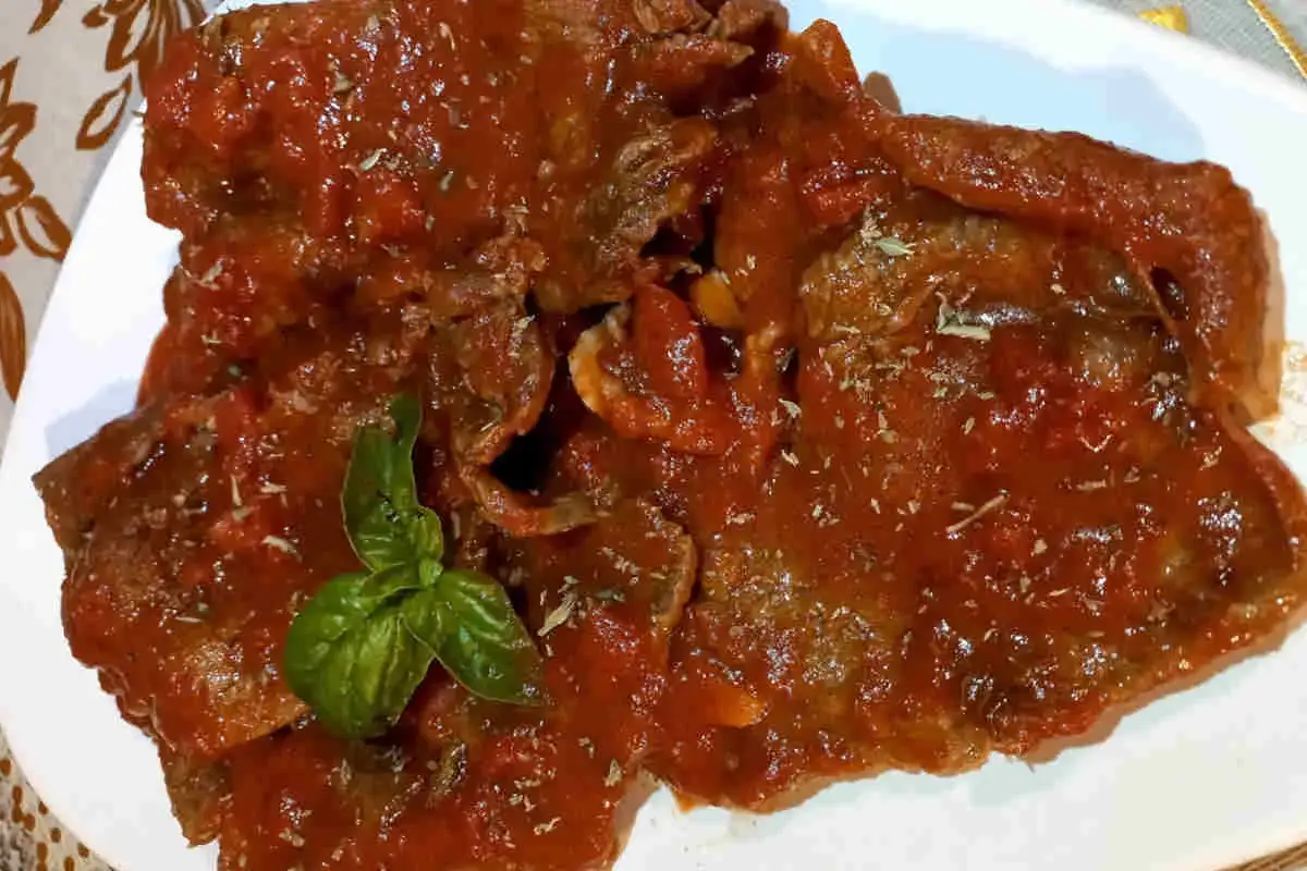 Un piatto invitante di carne alla pizzaiola morbida, ricoperta da un sugo rosso e profumato, guarnita con basilico fresco.