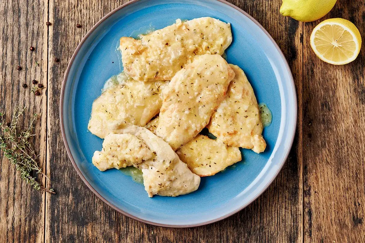 Piatto di pollo al limone con salsa cremosa, guarnito con erbe aromatiche e pepe nero su un piatto blu.