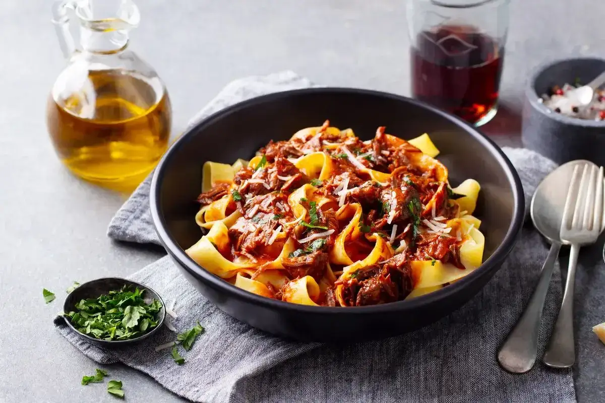 Pappardelle al cinghiale, un piatto ricco e saporito, servito in una ciotola scura con contorno di prezzemolo tritato e un bicchiere di vino rosso.