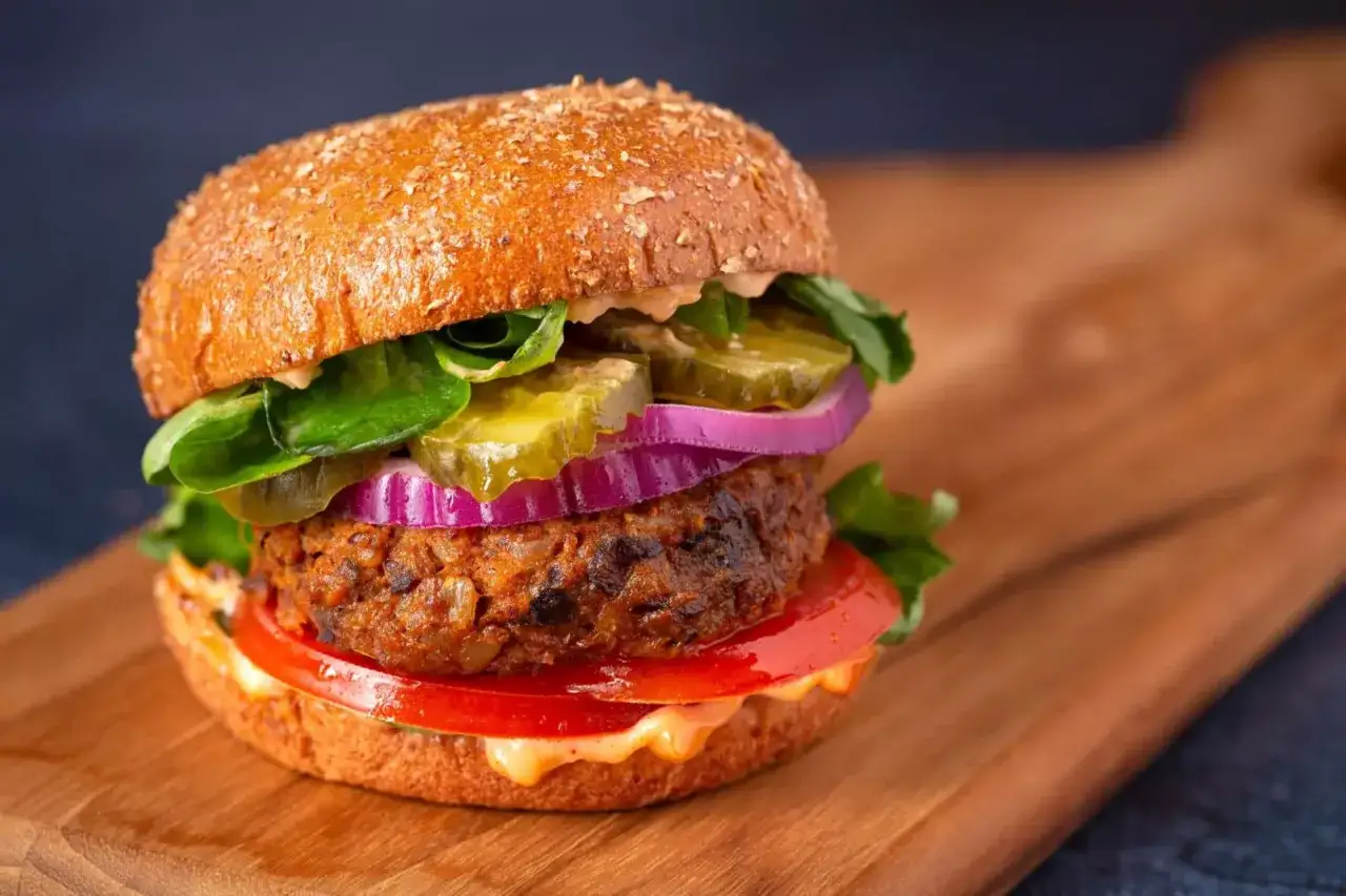 Hamburger di melanzane con pomodoro, cipolla rossa, cetriolini e lattuga su tagliere di legno.