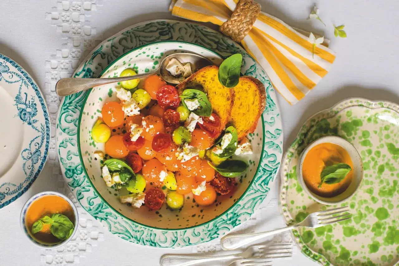 Insalata estiva con melone, pomodorini, feta e basilico, servita con pane tostato e una piccola ciotola di gazpacho.