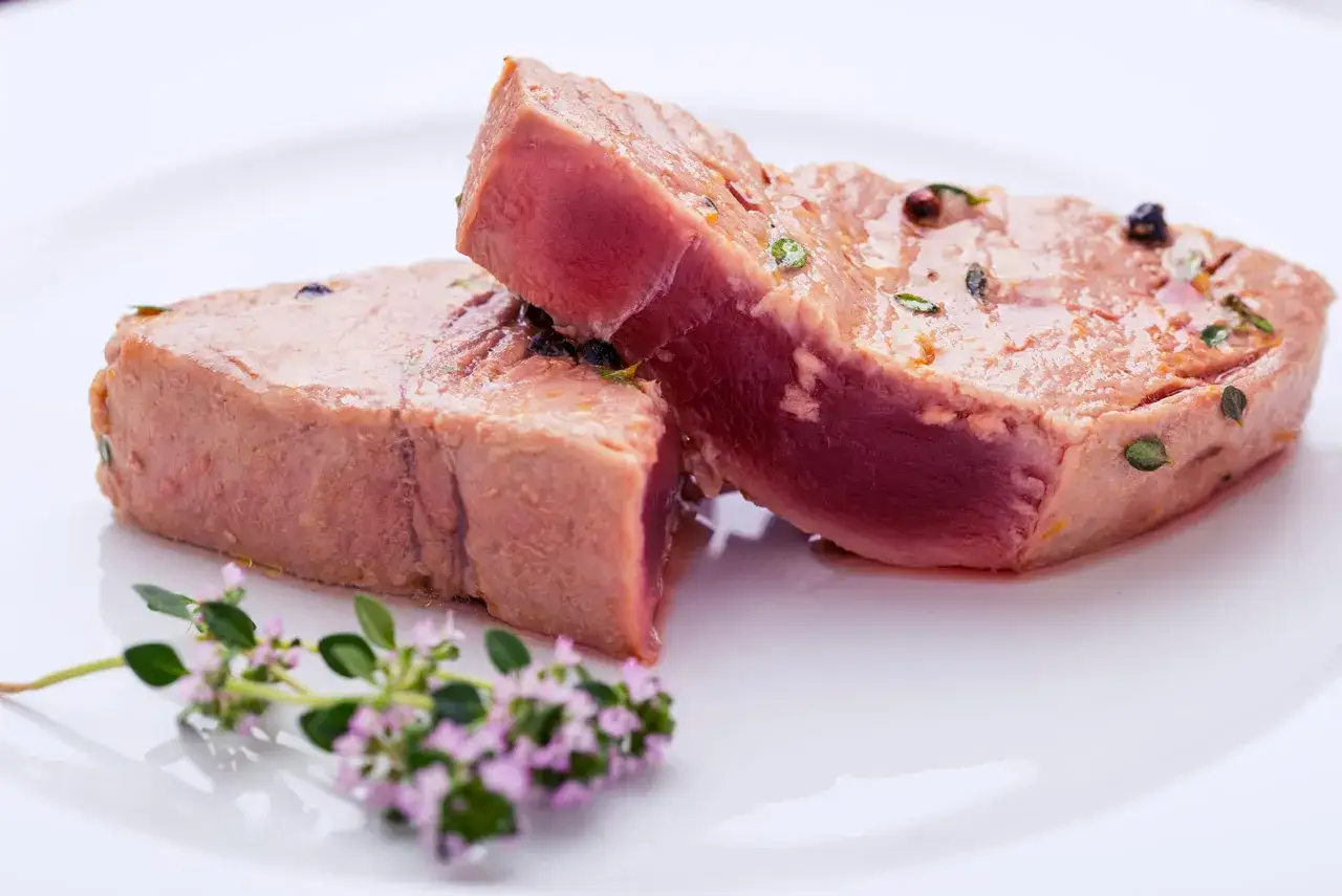 Due tranci di tonno scottati, conditi con pepe e erbe aromatiche, su un piatto bianco con un rametto di fiori viola.