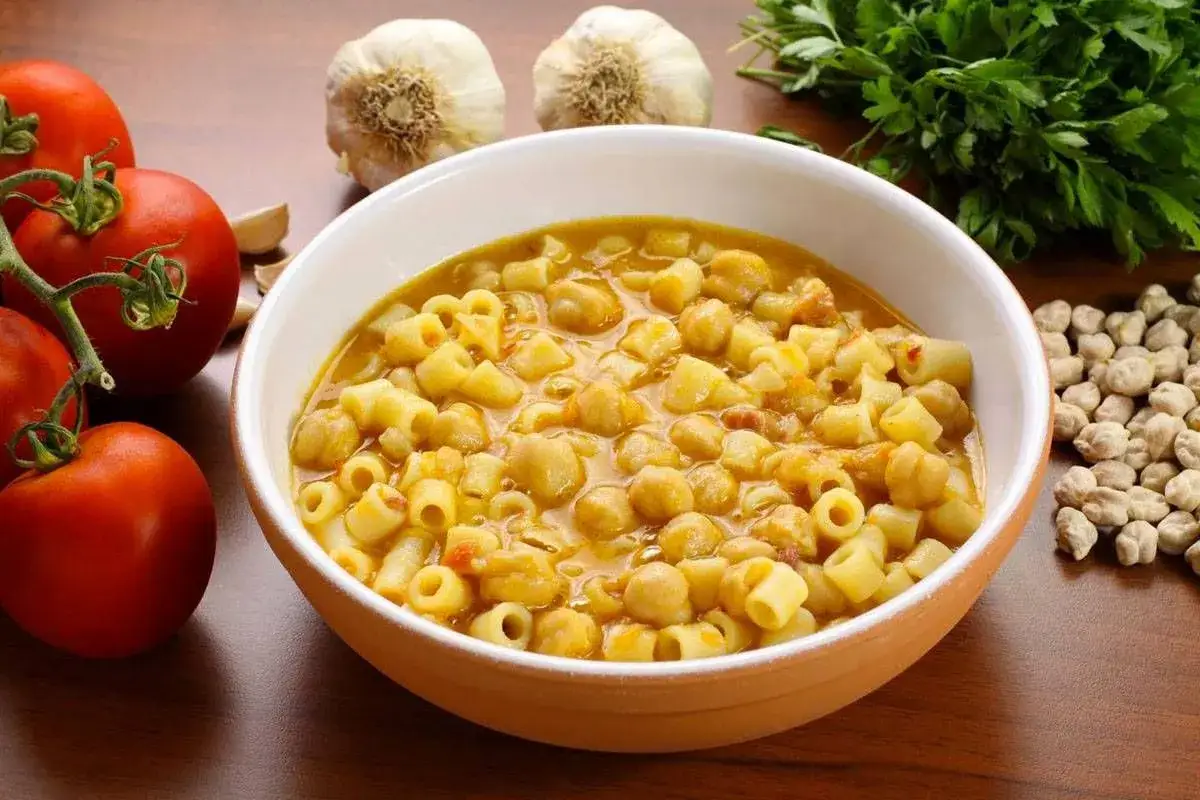 Un piatto fumante di pasta e ceci, con pomodori, aglio e prezzemolo sullo sfondo.