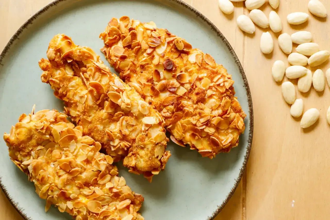 Tre cotolette di pollo impanate con mandorle a lamelle, dorate e croccanti, servite su un piatto azzurro.