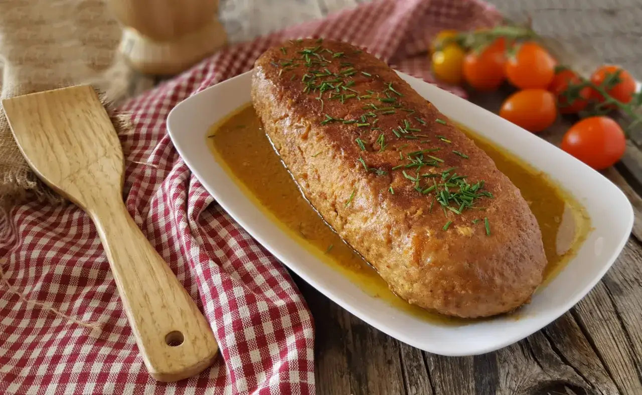 Polpettone in padella dorato e succulento, guarnito con erba cipollina, servito su un piatto bianco con salsa. Accanto, una spatola di legno e pomodorini.