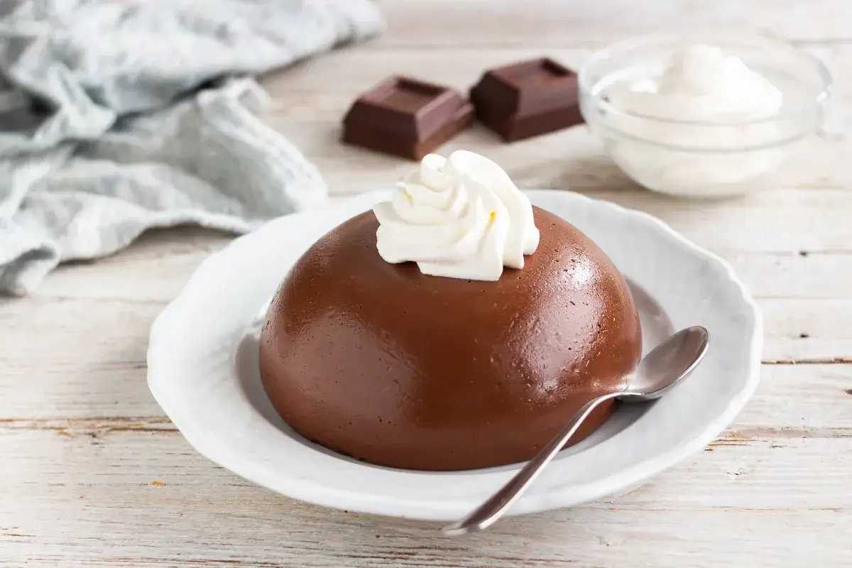 Budino al cioccolato liscio e lucido, guarnito con panna montata, su un piatto bianco. Accanto, due quadratini di cioccolato e una ciotola di panna.