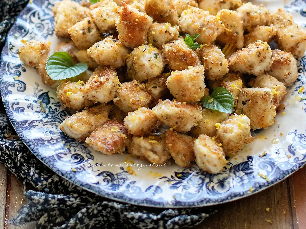 Bocconcini di pollo dorati e croccanti, guarniti con scorza di limone e foglie di menta fresca, serviti su un piatto decorato.