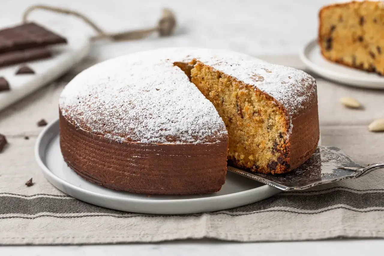Deliziosa torta con farina di mandorle, cosparsa di zucchero a velo, con una fetta sollevata da una paletta d'argento. Pezzi di cioccolato e mandorle sullo sfondo.