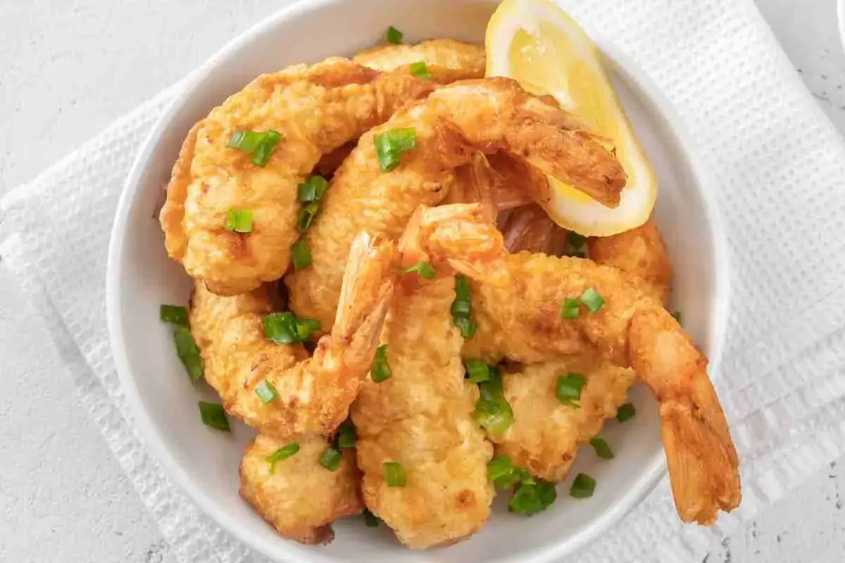Gamberi in tempura dorati e croccanti, guarniti con erba cipollina e una fetta di limone, serviti in una ciotola bianca.