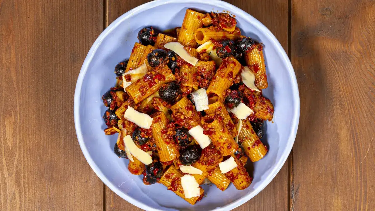 Rigatoni con nduja, olive nere e scaglie di parmigiano in una ciotola blu su un tavolo di legno.