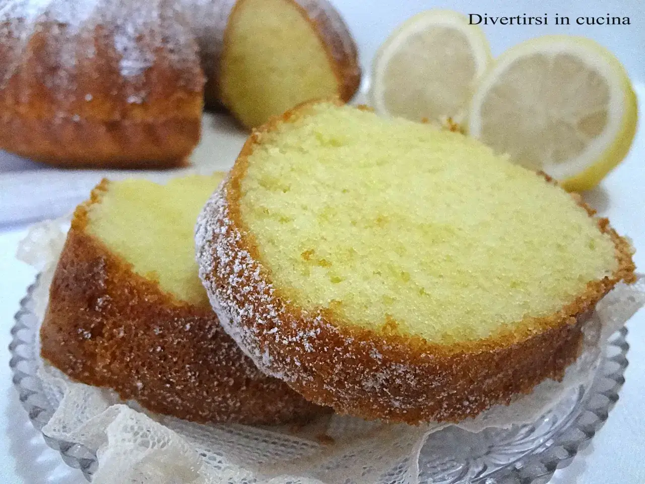Fette di torta al limone velocissima, spolverate di zucchero a velo, con fette di limone sullo sfondo.