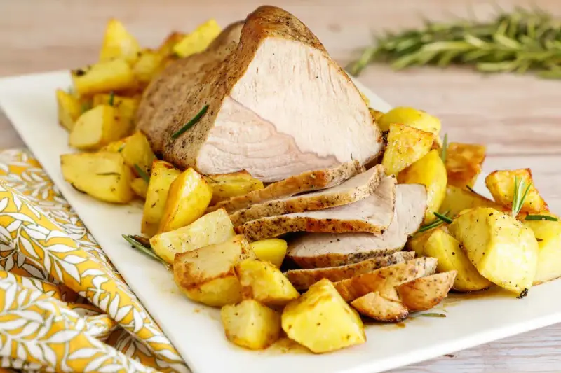 Un succulento arrosto di vitello affettato, servito su un piatto bianco con patate arrosto dorate e rametti di rosmarino.